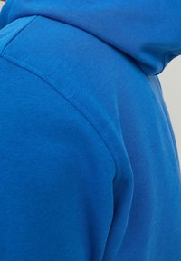 Detailní záběr na modrou tkaninu, znázorňující ramenní šev a rukáv oděvu s hladkou strukturou a pečlivým šitím.