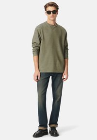 Junger Mann mit schwarzen Sonnenbrillen, olivgrünem Langarmshirt, dunklen Jeans mit Umschlägen und schwarzen Schuhen, der vor einem weißen Hintergrund steht.