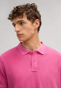 Roze poloshirt met kraag, korte mouwen en een twee-knoop sluiting aan de voorkant. Heeft een gestructureerde stof en een klein logo op de borst.