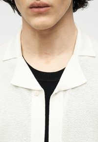 Άνδρας που φοράει λευκό πουκάμισο με υφή και κουμπιά πάνω από μαύρο πλεκτό πουλόβερ, εμφανιζόμενο από τον λαιμό μέχρι το πάνω μέρος του στήθους, με φόντο ένα απλό φόντο.