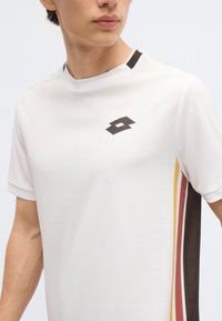 Νεαρό άτομο που φορά λευκό αθλητικό πουκάμισο με κοντά μανίκια, με μαύρο λογότυπο στο στήθος και κάθετες κίτρινες, κόκκινες και μαύρες ρίγες στο πλάι.