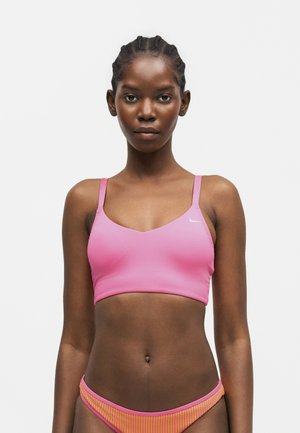 Kvinde med flettet hår iført en pink sports-bh og orange bikiniunderdel mod en ensfarvet hvid baggrund.