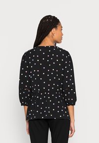 Blusa preta com bolinhas coloridas, mangas 3/4 e design peplum. Apresenta um delicado acabamento em renda nas aberturas das mangas. Textura de tecido suave.