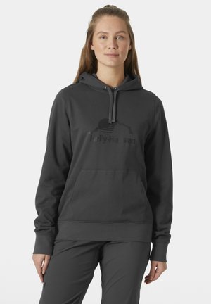 Vrouw in een donkergrijze Helly Hansen-hoodie en bijpassende broek, staand tegen een effen lichte achtergrond, naar voren kijkend.