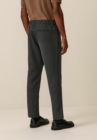 Pantalons ajustés gris charbon avec une ceinture élastique, coupe slim, texture lisse et détail de poche arrière unique. Portés avec des chaussures noires.