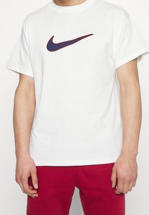 Personne portant un T-shirt blanc avec un logo swoosh brodé bleu foncé et bordeaux, et un pantalon rouge, debout devant un fond uni.