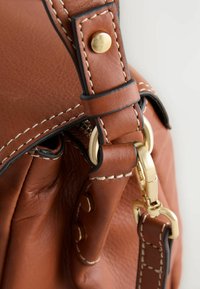 Dettaglio di una borsa a mano in pelle marrone che mostra una chiusura in metallo dorato, cuciture a contrasto e texture in tessuto sui manici e sulla borsa, con chiusura a zip.