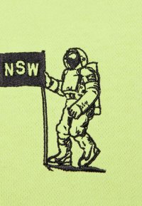 Broderi af en astronaut i en sort dragt, der holder et flag med "NSW," sat mod en lys gul stofbaggrund.