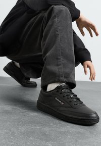 Sorte skinnesneakers med teksturert gummisåle, snøring, polstret krage og Reebok-logo på siden, plassert mot grått gulv.