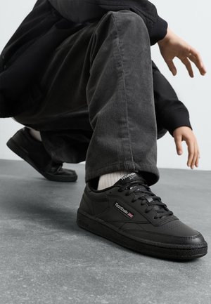 Sapatilhas de couro preto com sola de borracha texturizada, design de atacadores, colarinho almofadado e logotipo da Reebok na lateral, colocadas sobre um chão cinza.