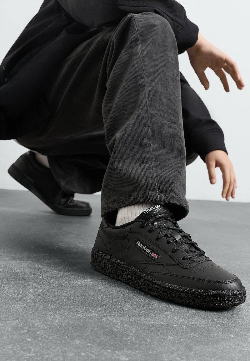 Sorte skinnesneakers med teksturert gummisåle, snøring, polstret krage og Reebok-logo på siden, plassert mot grått gulv.