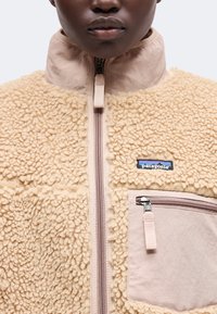 Κοντινό πλάνο ενός ατόμου που φοράει μπεζ fleece μπουφάν Patagonia με τσέπη στο στήθος με φερμουάρ και ψηλό γιακά.