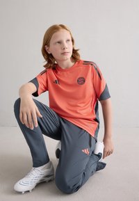 Conjunto desportivo juvenil com uma t-shirt corais vibrantes com detalhes em cinza e logótipo, combinada com calças cinza escuras e chuteiras brancas.