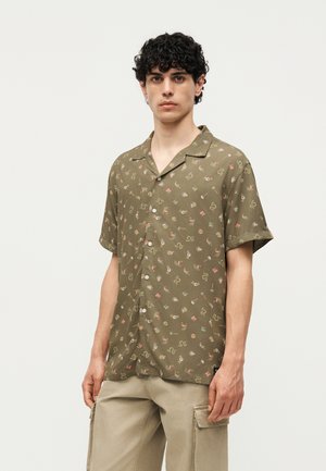 Młody mężczyzna z kręconymi ciemnymi włosami, w oliwkowo-zielonej koszuli na krótki rękaw z małymi kolorowymi wzorami i beżowych spodniach typu cargo.