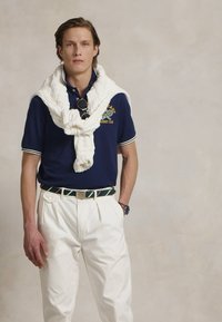 Navy polo with white accents, knitted white sweater draped around neck, white pleated trousers, green and white striped belt, sunglasses.