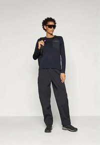 Persona con camiseta de manga larga negra, pantalones oscuros holgados, zapatos negros, gafas de sol y llevando una correa de mochila negra sobre un hombro.