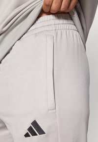 Ανοιχτό γκρι παντελόνι φόρμας με ελα στική μέση, πλαϊνές τσέπες και μαύρο λογότυπο Adidas στο αριστερό πόδι. Μαλακή, λεία υφή υφάσματος.
