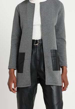 Cardigan gris texturé ouvert devant avec poches en cuir noir porté sur un haut blanc côtelé et un pantalon en cuir noir avec ceinture à boucle argentée.