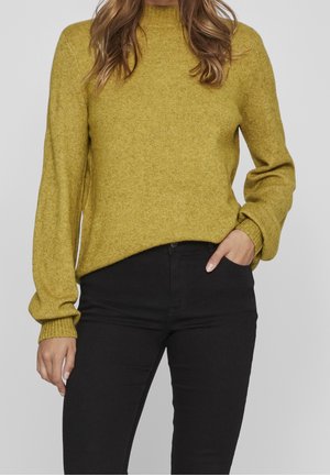 Frau trägt einen senfgelben Strickpullover mit langen Ärmeln und schwarze eng anliegende Hose, steht mit einer Hand in der Tasche vor einem einfarbigen Hintergrund.