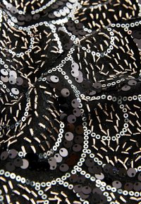 Čierna tkanina ozdobená kruhovými flitrami a kovovými tyčinkami usporiadanými do vírových vzorov, vytvárajúc textúrovaný a odrážajúci povrch.