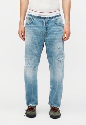 Mand iført lyseblå slidte jeans med falmede pletter, kombineret med grå og brune sneakers og en hvid linning synlig over jeansene.