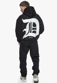 Schwarzer Hoodie mit großem weißen Grafikdesign auf dem Rücken, das stilisierte Buchstaben zeigt. Kombiniert mit schwarzen Hosen und grauen Sneakers. Lässiger Schnitt.