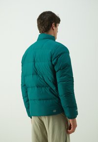 Teal pufferjakke med høj krave, quiltet tekstur og elastiske manchetter. Har en logodetalje nær kanten. Vist fra ryggen.