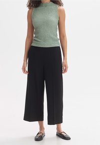 Světlounce zelený texturovaný top bez rukávů s vysokým límcem, doplněný černými širokými kalhotami a černými plochými botami.