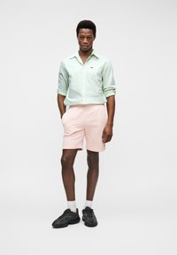 Homme debout les mains dans les poches, portant une chemise boutonnée vert clair, un short rose, des chaussettes blanches et des baskets noires sur fond blanc.