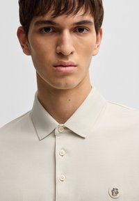 Beige polo skjorte med en myk tekstur, krage, fire knapper og en logo aksent på venstre bryst. Enkel og minimalistisk design.