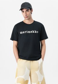 Fekete püffös pamut póló fehér "marimekko" logóval, kerek nyakkivágással, rövid ujjakkal, bézs mintás rövidnadrággal párosítva. Fekete sapka teszi teljessé a megjelenést.