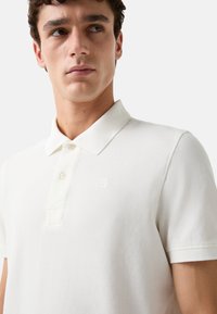 Hvid polo t-shirt lavet af struktureret stof, der har en tre-knaps knaplukning og et lille broderet logo på venstre bryst.