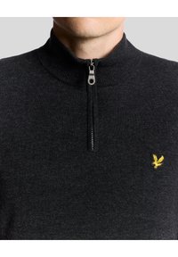 Svart stickad tröja med hög krage och fyrdelad dragkedja. Har ett litet gult örn-emblem på vänster bröst.