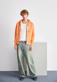 Narancssárga zipzáras kapucnis pulóver, elülső zsebekkel és logóval. Fehér ujjatlan póló alatta. Laza, világoskék farmernadrág. Szürke cipők. Egyszerű háttér.