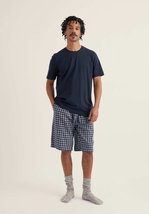 Hombre de pie con camiseta azul marino, pantalones cortos de cuadros azules y blancos, y calcetines grises contra un fondo claro y liso.