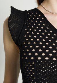 Gros plan d'une personne portant un haut noir tricoté avec un motif à tissage ouvert et un collier de perles argentées.