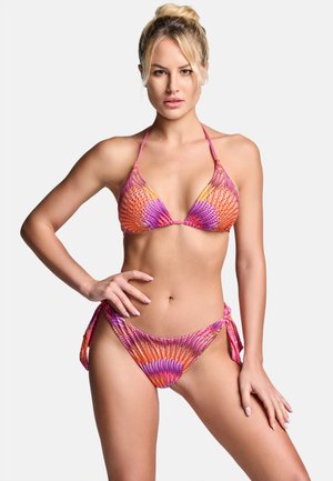 Kobieta o blond włosach w kolorowym pomarańczowo-różowym bikini w wzory, pozująca jedną ręką na biodrze na jednolitym jasnym tle.