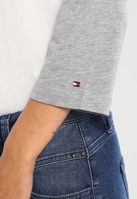 Graues Hemd mit drei Viertelärmeln und weißem Körper, versehen mit einem kleinen gestickten Logo in Marineblau, Rot und Weiß am Bund. Jeans aus Denim mit Akzenten.