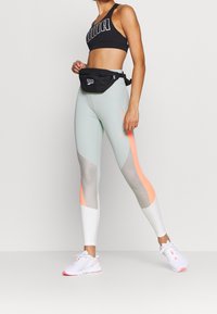 Sujetador deportivo negro corto con un logotipo destacado, leggings menta y blancos con acentos naranjas y paneles de malla, complementados con zapatillas deportivas blancas.