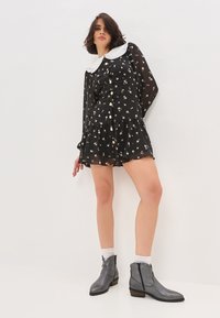 Vestido mini negro con estampado floral, mangas largas transparentes, cuello Peter Pan blanco y botones dorados. Llevado con botas cortas grises y calcetines blancos.
