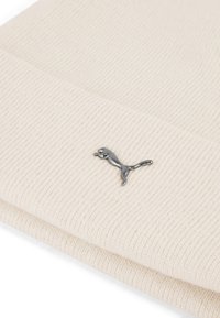 Bonnet en tricot beige avec une texture côtelée, arborant un petit logo métallique argenté à l'avant. Matériau doux et flexible.
