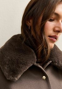 Bruine jas met een bontkraag, met een zachte textuur, gouden knoop en een getailleerd ontwerp. Close-up van de kraag en zijaanzicht van het gezicht.