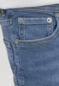 Zili denim džinsi ir piecu kabatu dizains, ar mazu priekšējo kabatu un metāla kniedēm, kas akcentē šuves. Faktūra ir gluda, ar redzamām dūrēm.