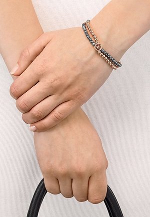 Deux mains serrées, un poignet portant deux bracelets à perles en or rose et gris foncé, tenant une poignée de sac noire sur un fond blanc texturé.