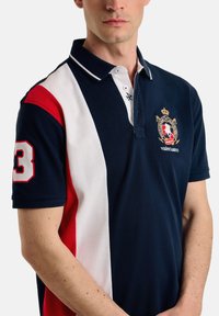 Polo de color navy con paneles rojos y blancos, mangas cortas, collar de dos botones y escudo bordado en el pecho. Presenta un gran número "3".