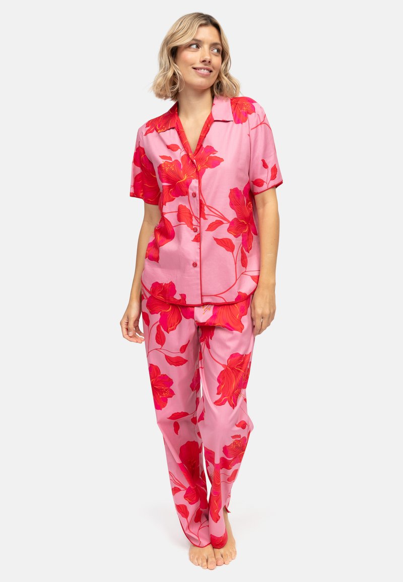 Kobieta ubrana w różowy satynowy komplet piżamowy w kwiaty z dużymi czerwonymi kwiatowymi wzorami, stojąca boso, uśmiechnięta i patrząca na bok.
