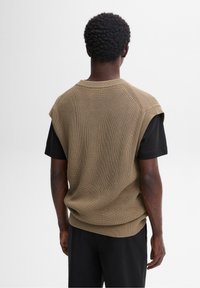 Man with short curly hair wearing a beige knitted sleeveless sweater over a black short-sleeve shirt, facing away on a white background.