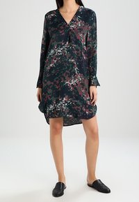 Vestido de manga larga en verde oscuro con patrones abstractos en burdeos, rosa y blanco. Escote en V y corte holgado con un dobladillo trasero más largo.