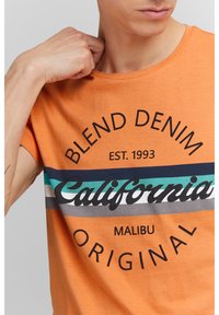 Narancssárga pamut póló fekete és többszínű csíkokkal díszített elülső grafikával, amelyen "California" felirat látható kör alakú dizájnnal és szöveges kiegészítésekkel.