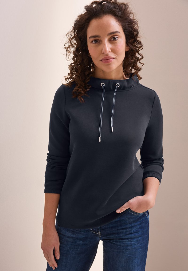 Dunkelgrauer Sweatshirt mit hohem Kragen, Kordelzugakzenten und langen Ärmeln, kombiniert mit blauen Jeans. Glatte Textur und schlankes Design.